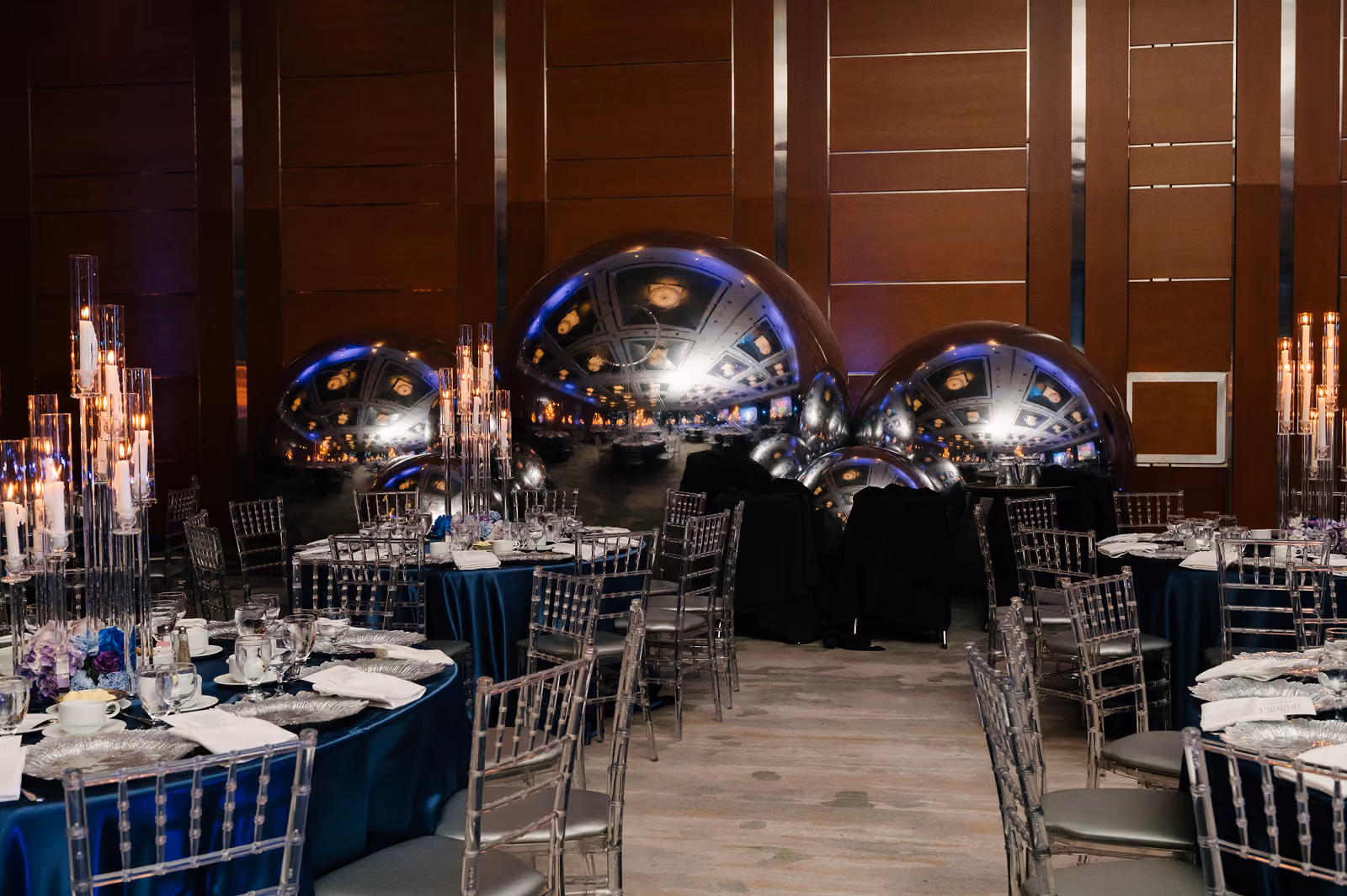 Event space with round tables, clear chairs, tall candle centerpieces, and large reflective silver spheres.