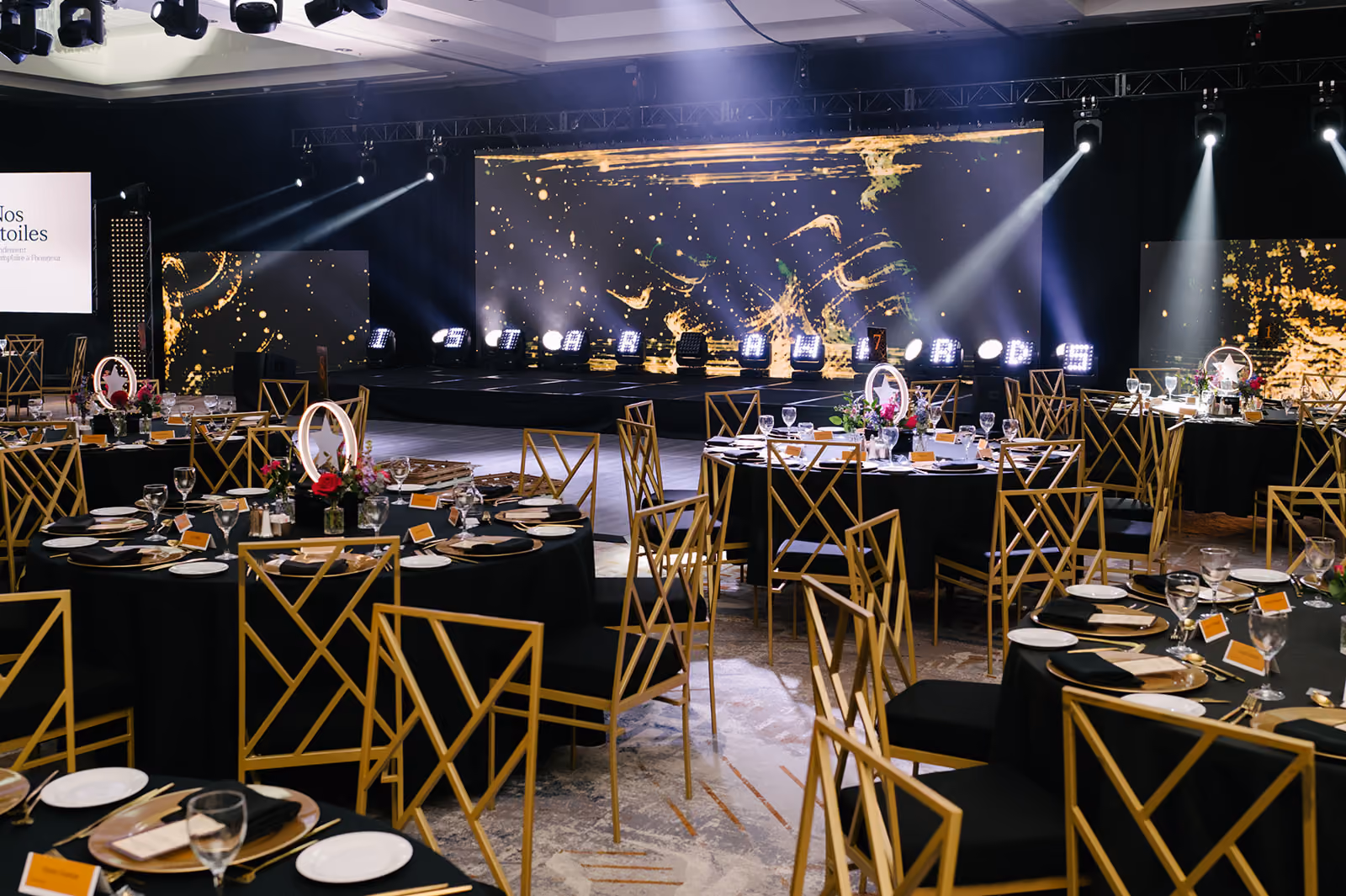 Elegant event space with round tables set for dinner and a lit stage with abstract golden designs.