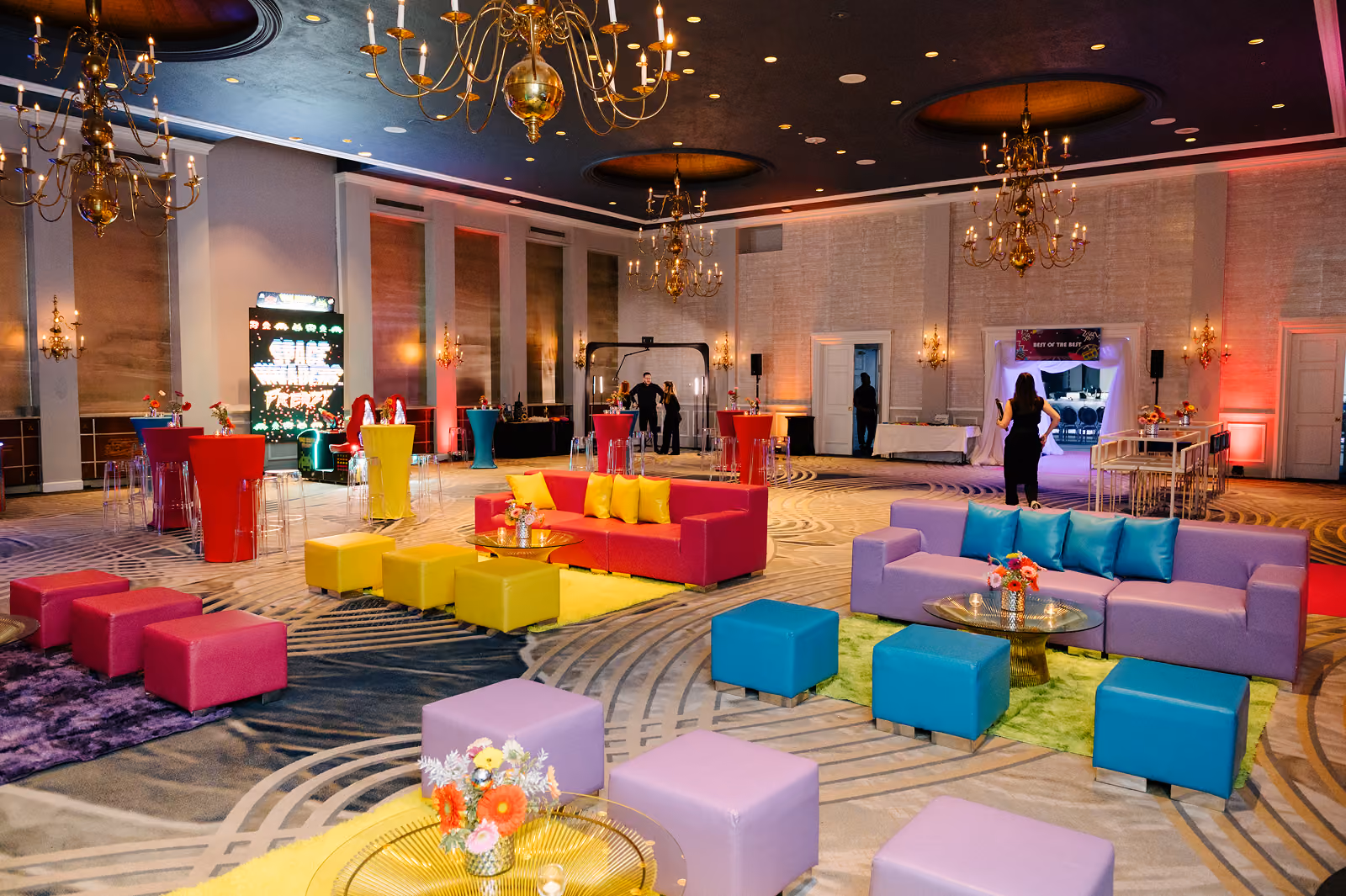 Colorful lounge area with sofas, ottomans, and chandeliers in a spacious event hall with people.