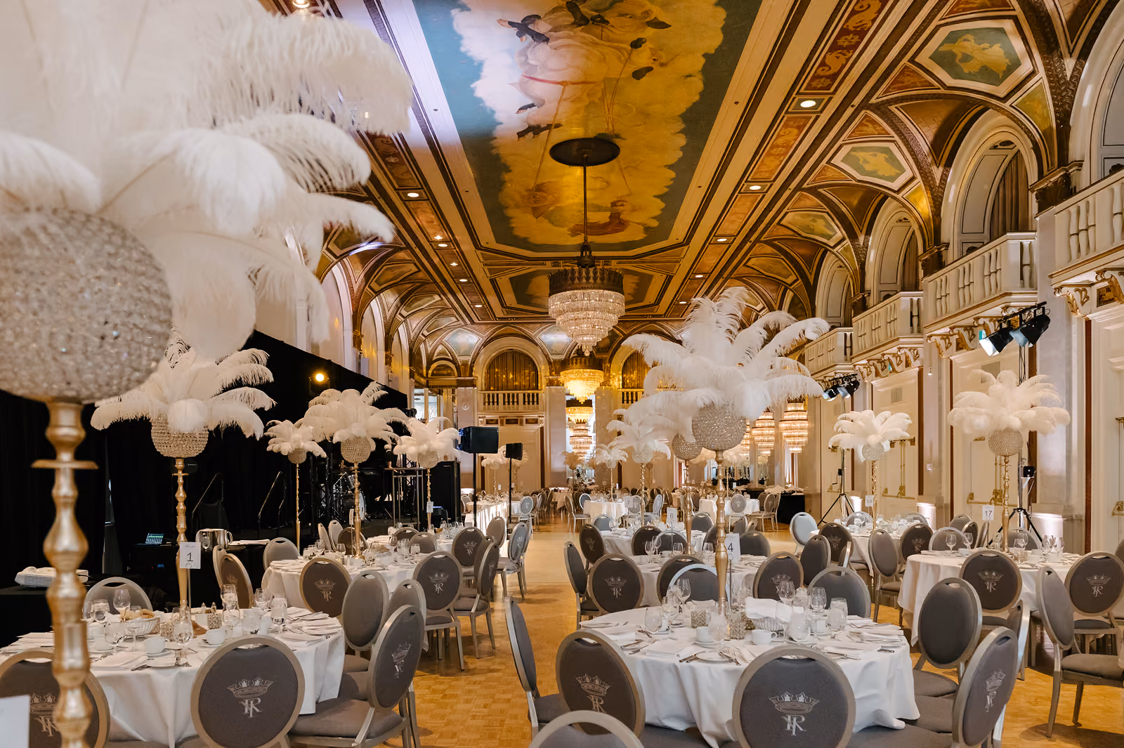 Elegant banquet hall with round tables, grey chairs, white feather centerpieces, and ornate painted ceiling.