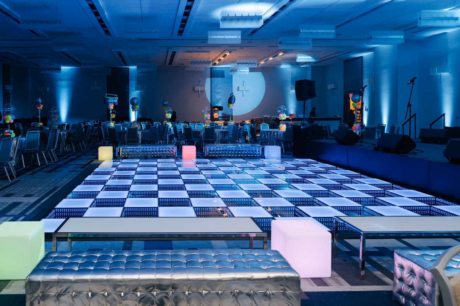 Indoor event space with LED chessboard dance floor, balloons, tables, silver benches, and stage with microphones.