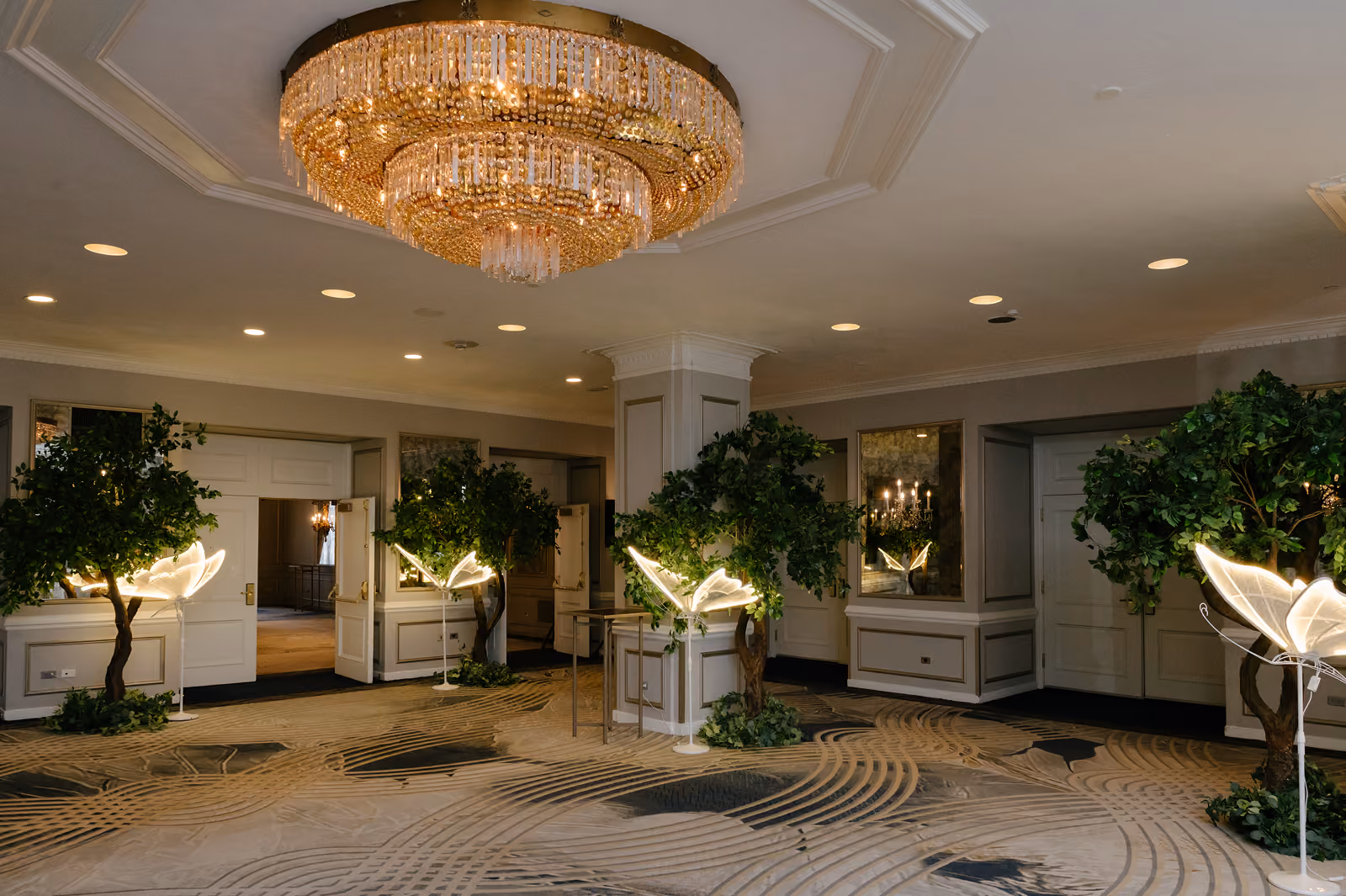 Luxurious room with a grand chandelier, potted trees, and glowing butterfly lights near open doors.