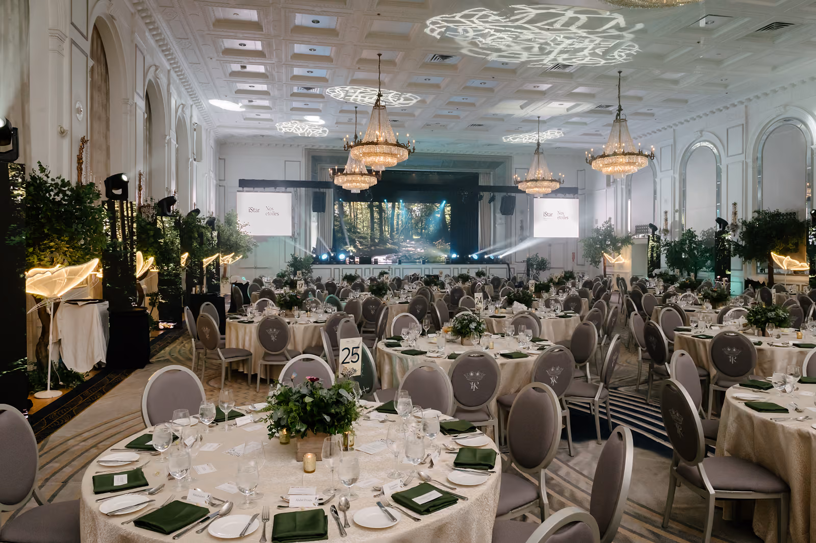 Elegant banquet hall with round tables set for dinner, chandeliers, and a stage with lighting effects.