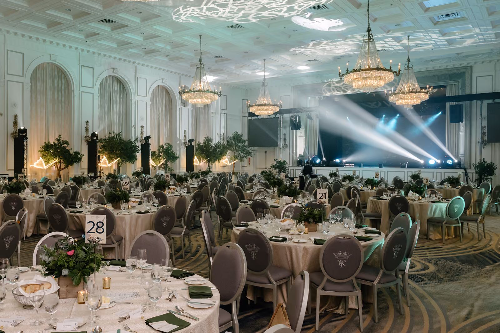 Elegant banquet hall with round tables set for event, chandeliers, stage, and green floral centerpieces.