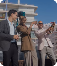 Three diverse adults dressed in business casual attire celebrating outdoors on a rooftop with a clear blue sky and apartment buildings in the background, smiling and opening a bottle of champagne.
