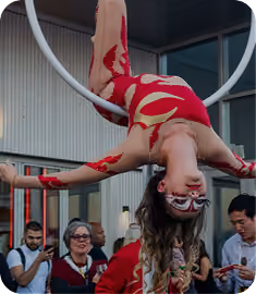 A woman wearing a red and beige costume with body paint performs aerial acrobatics on a white hoop, hanging upside down with arms extended, while a small crowd watches in the background.