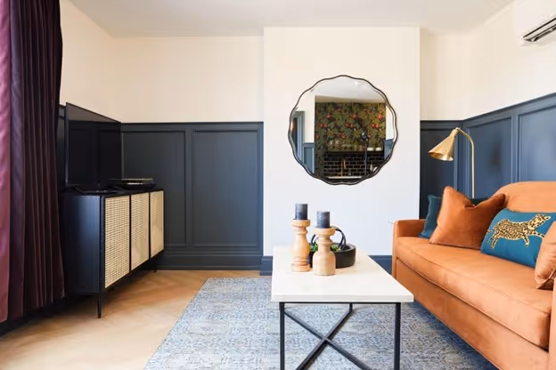 This chic living room features a warm orange sofa, a marble-topped coffee table, and bold wall paneling in dark tones. Accented with modern décor and a stylish round mirror, the space offers a blend of comfort and contemporary design.