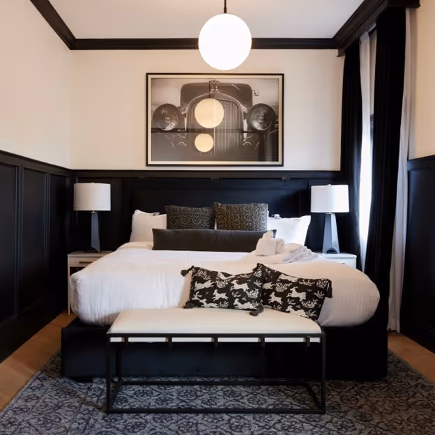 This elegantly designed guest room features a cozy, plush bed framed by modern black wainscoting, accented with stylish lighting and artwork. The inviting space combines classic and contemporary touches for a luxurious and comfortable stay