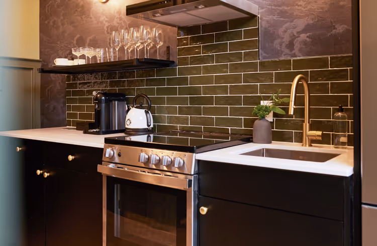 The Dorchester's rooms feature modern kitchenettes with sleek black cabinetry, stainless steel appliances, and a stylish green subway tile backsplash
