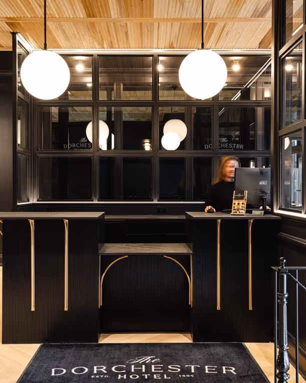 The lobby entrance of The Dorchester Hotel welcomes guests with a sleek, modern design featuring bold black finishes, elegant lighting, and warm wood accents. This stylish space sets the tone for a luxurious and refined guest experience
