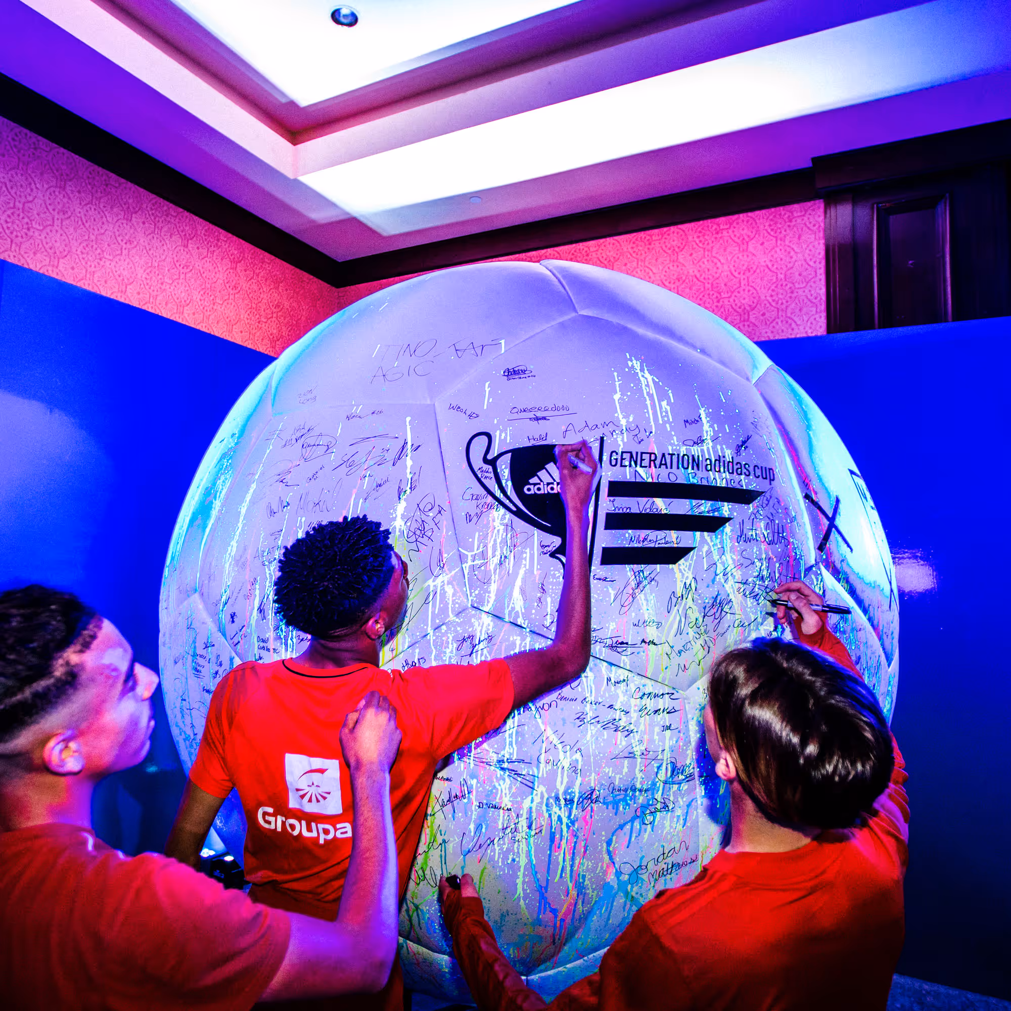 Three young men in red athletic shirts are signing a massive, glowing white soccer ball at an indoor event. The ball features the Generation adidas cup logo and a trophy illustration, and it is covered in handwritten signatures. The room is dimly lit with vibrant purple and blue ambient lighting