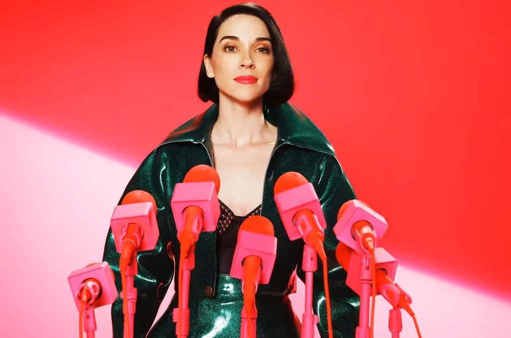 An eye-catching, high-contrast portrait of the artist St. Vincent (Annie Clark). She has short, dark hair and bold red lipstick, looking directly at the camera with a calm expression. She is wearing a metallic, dark green textured jacket over a black mesh top. A cluster of several bright pink microphones on matching pink stands is positioned in front of her, set against a vibrant, solid red background.