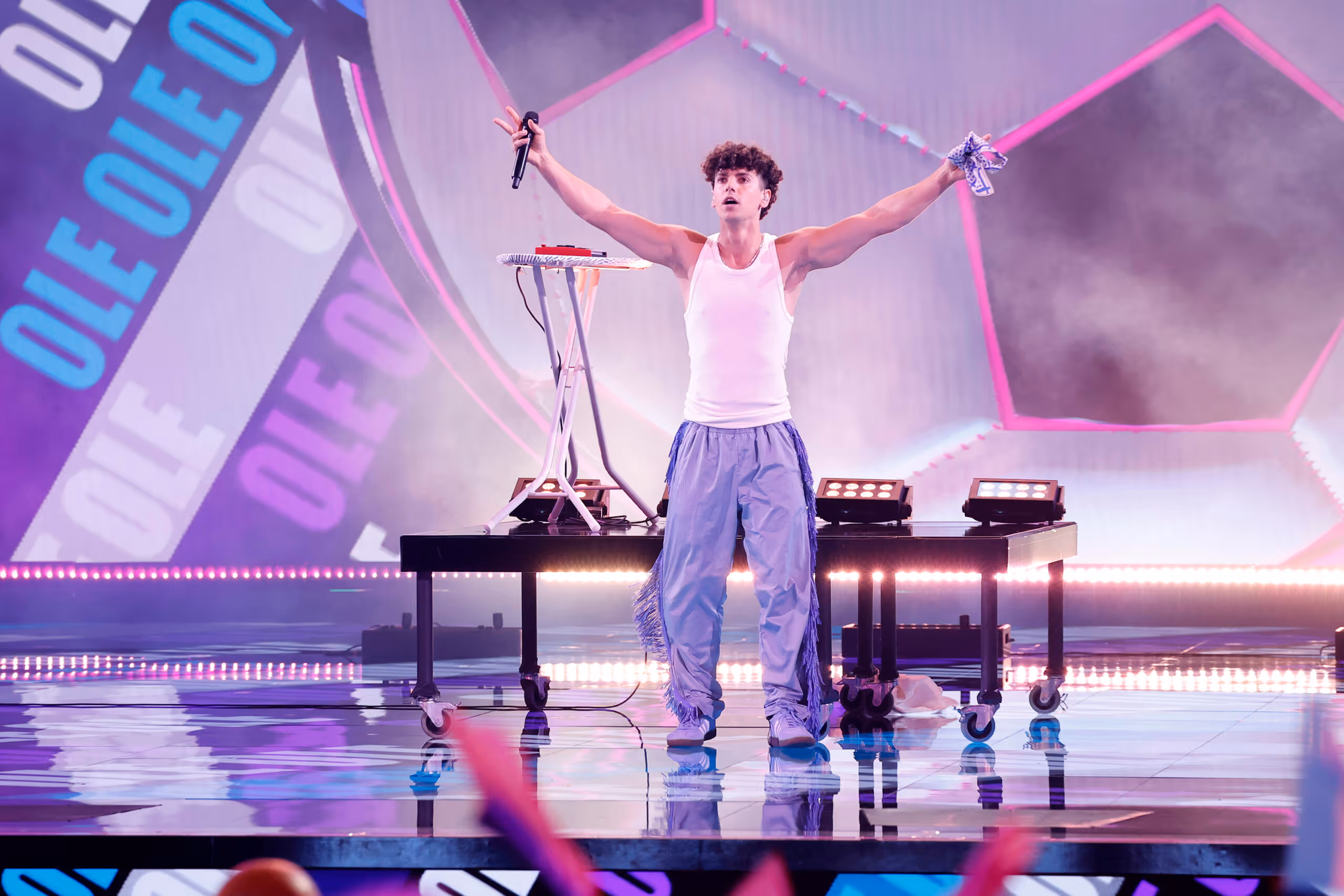 A high-energy stage performance featuring a young male singer with curly hair, wearing a white tank top and baggy light-purple trousers with fringe detailing. He stands behind a long table with his arms outstretched and head tilted back, holding a microphone in one hand. The background features vibrant pink lighting and large banners with "OLE" printed diagonally in blue and white.