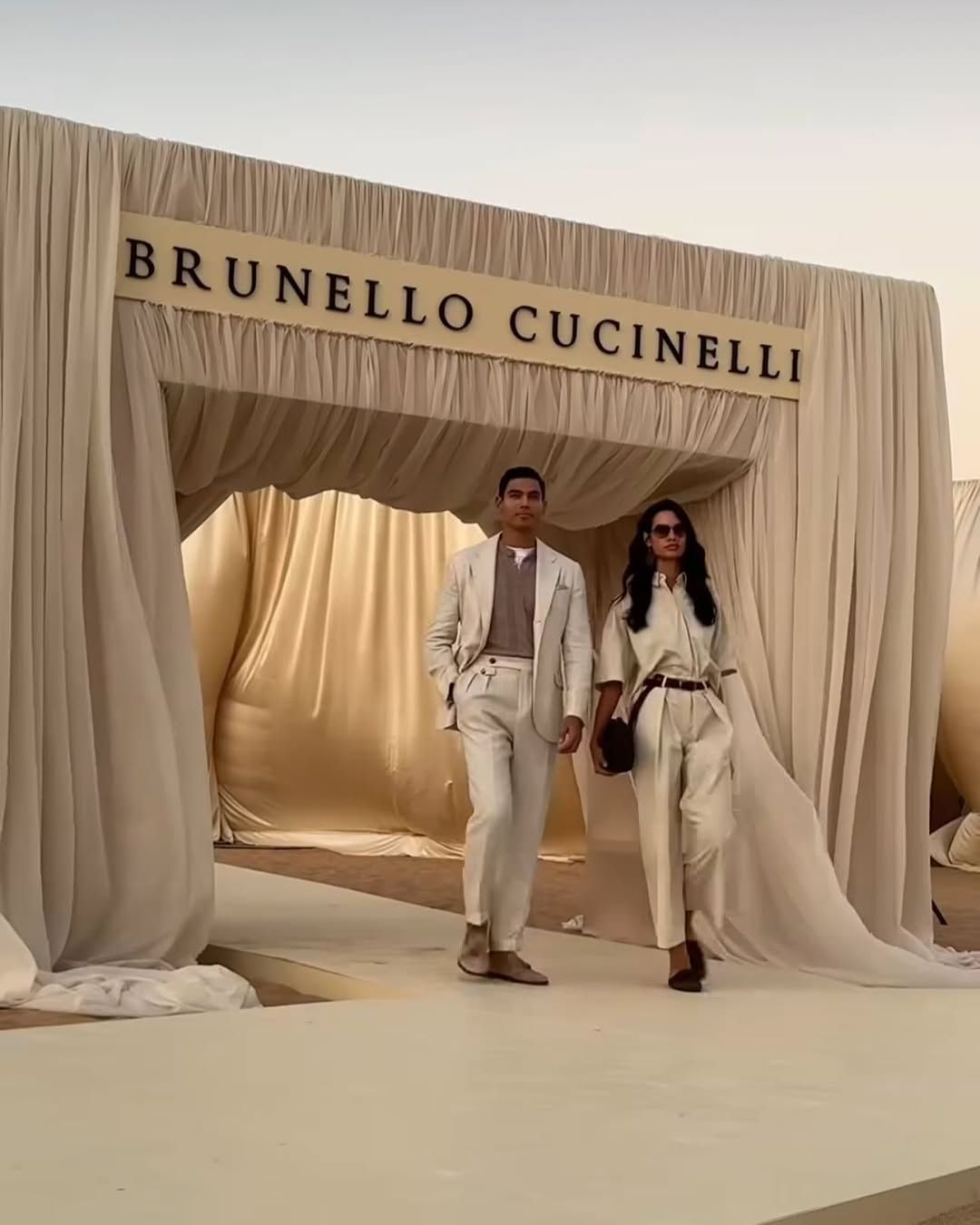 Two models in light-colored outfits walking under Brunello Cucinelli archway with flowing fabric backdrop