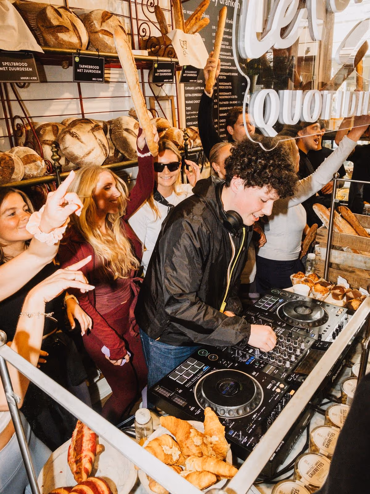 An upbeat, vertical photograph capturing a DJ performing at a lively event inside what appears to be a bakery or gourmet food shop.