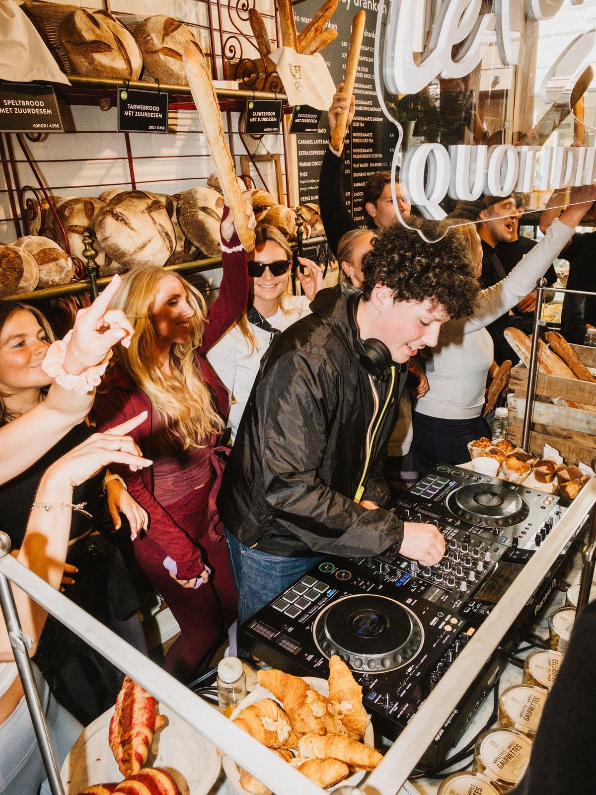 An upbeat, vertical photograph capturing a DJ performing at a lively event inside what appears to be a bakery or gourmet food shop.