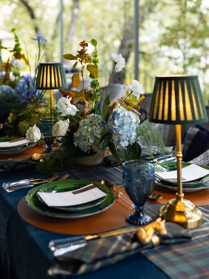 An elegant outdoor dinner table setting featuring green plates, gold-base lamps with pleated shades, and lush floral centerpieces with white and yellow flowers.