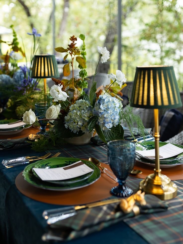 An elegant outdoor dinner table setting featuring green plates, gold-base lamps with pleated shades, and lush floral centerpieces with white and yellow flowers.