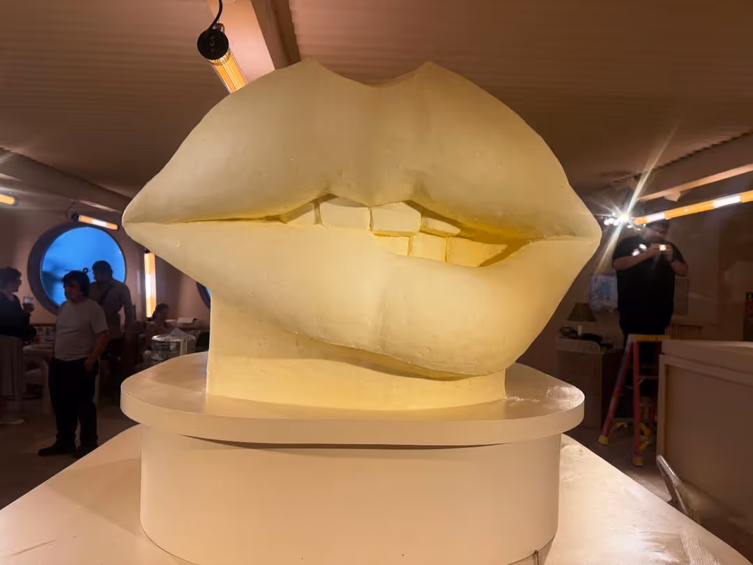 Large, pale yellow sculptural bust of a pair of lips and teeth resting on a white pedestal in an indoor event space with people in the background and "The Idea Hunter" logo in the top-left corner.
