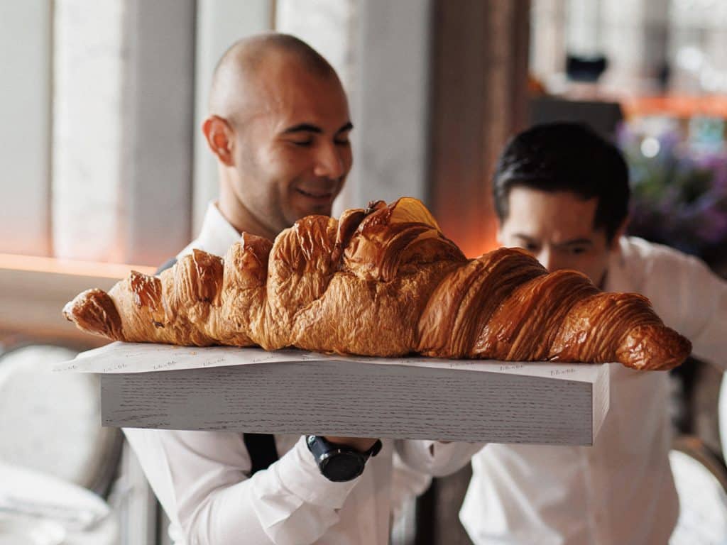 Two men in white chef coats holding a large wooden tray featuring an exceptionally oversized, golden-brown flaky croissant. The logo for "The Idea Hunter" is visible in the top left corner.