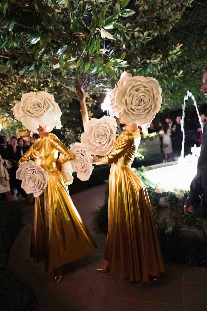Two performers wearing floor-length metallic gold gowns and large, oversized white rose headpieces that completely cover their faces, posing in a garden setting at night near a lighted fountain. The logo for "The Idea Hunter" is visible in the top left corner.