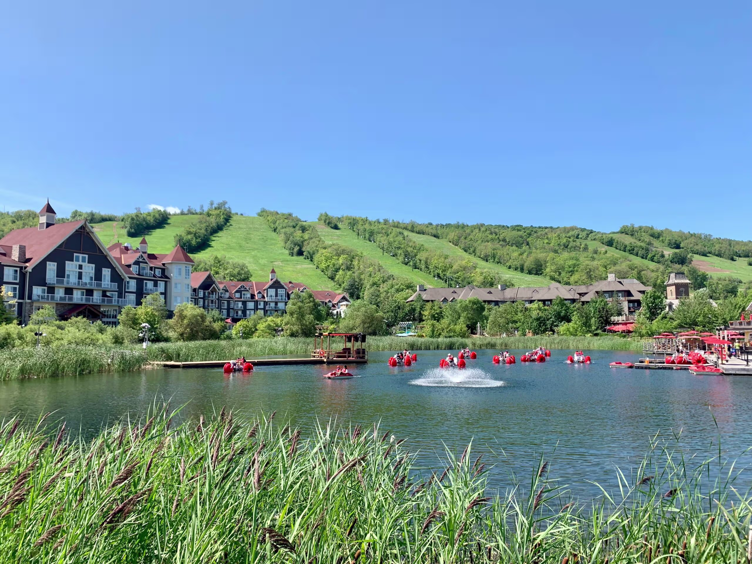 Scenic lakeside resort with red-roofed buildings, fountain, and paddle boats against green hills