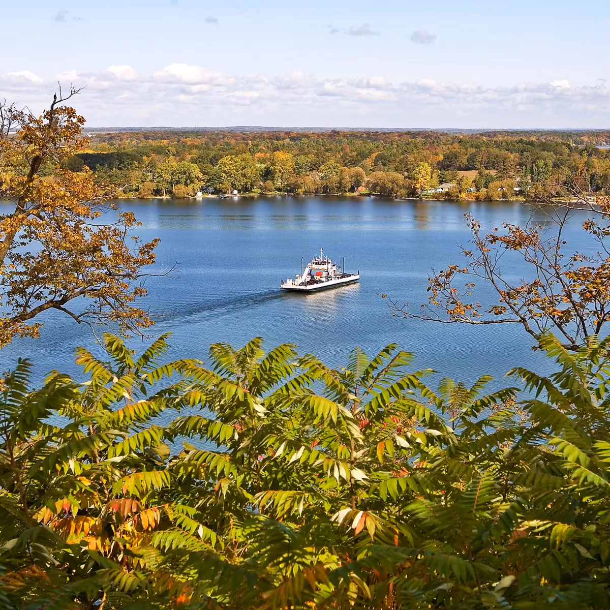 Ferry boat carrying vehicles across calm lake with autumn-colored trees and partly cloudy sky