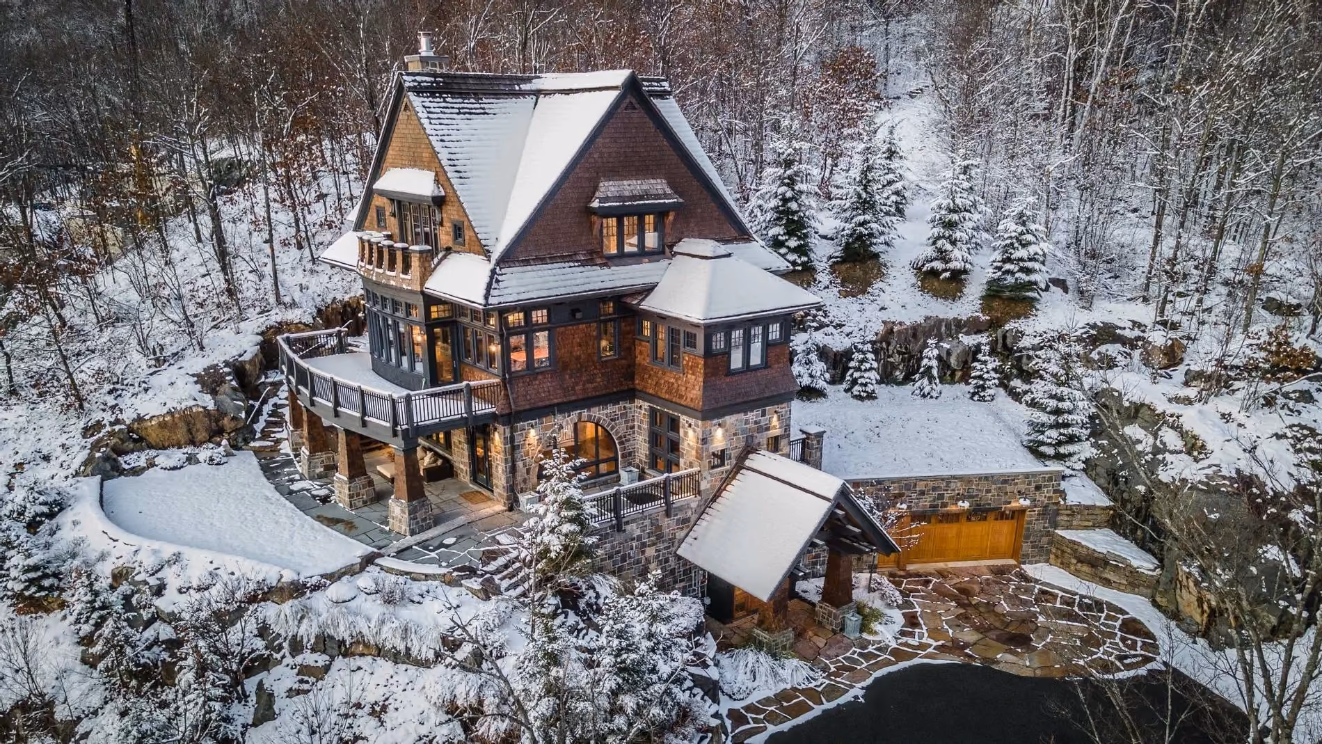 Large multi-story stone and wood house with snow-covered roof, warm lights, and snowy forest landscape