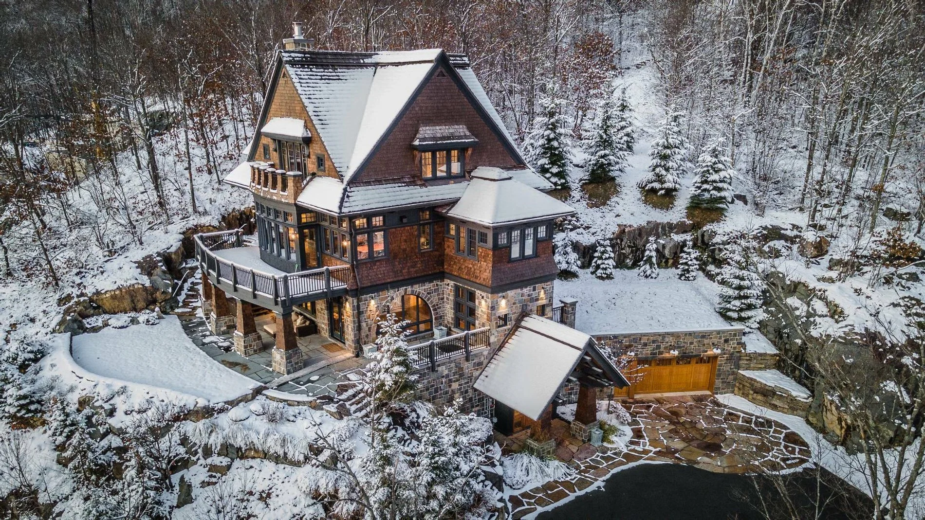 Large multi-story stone and wood house with snow-covered roof, warm lights, and snowy forest landscape