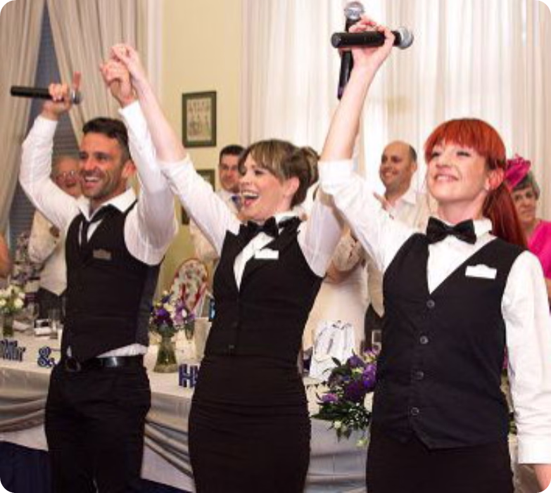 Three performers in formal attire singing with microphones at a decorated event table