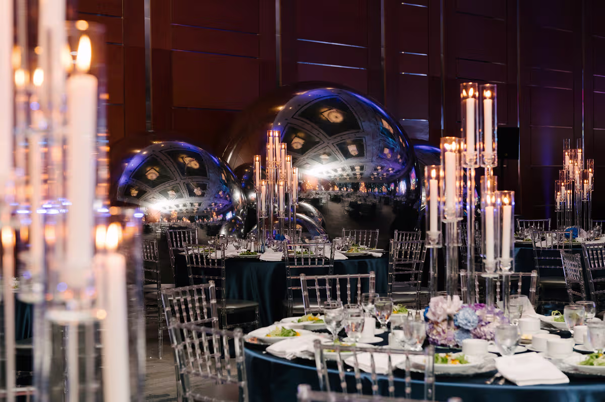 Banquet hall with round tables, tall candle holders, floral centerpieces, transparent chairs, and reflective silver spheres