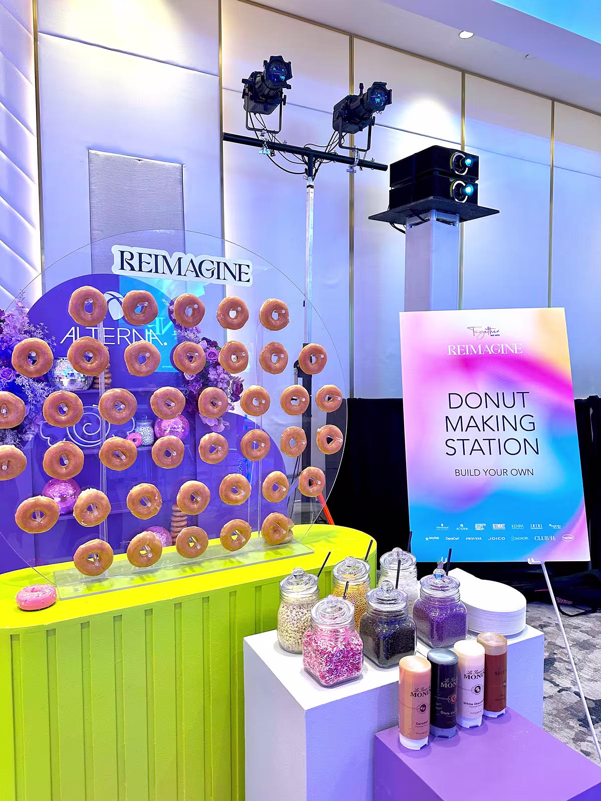 Colorful donut making station with glazed donuts on pegs, toppings jars, and Alterna Reimagine signage