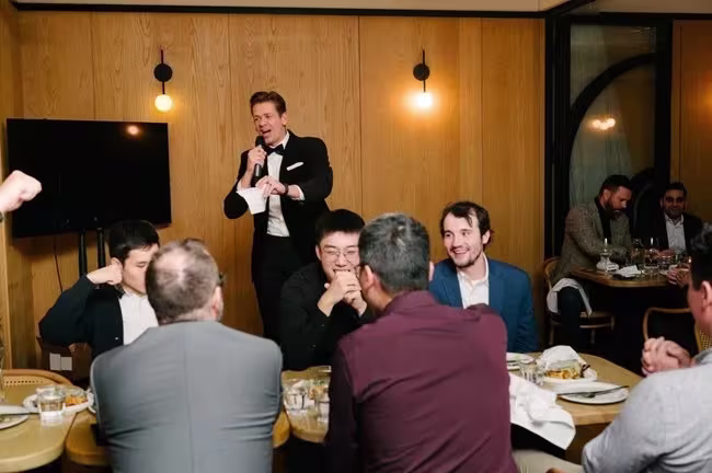 A host with a microphone and cue cards works the room at a seated holiday dinner.