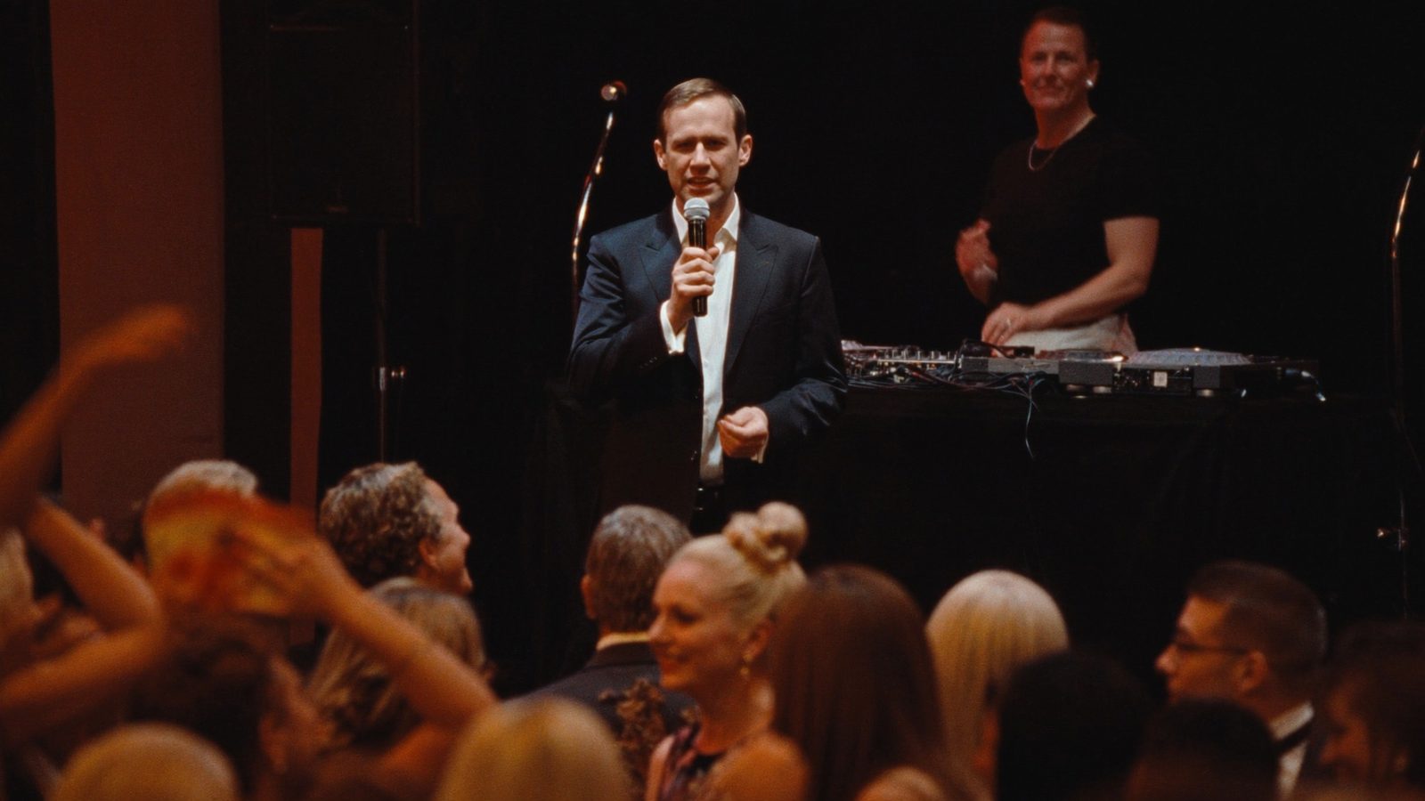 Emcee speaking on stage with a DJ at the booth behind him and guests on the dance floor