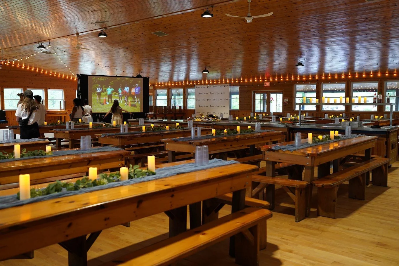 A camp dining hall set for a retreat dinner with candles, greenery and string lights.