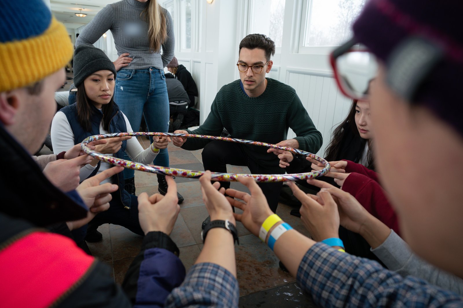 A team crouches in a circle, each person holding a hoop on their fingertips during a team challenge