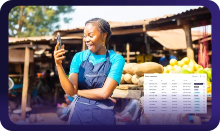Smiling woman in blue apron looking at smartphone at an outdoor market with root vegetables and lemons on display.