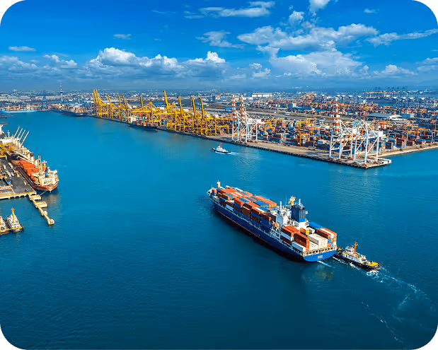 Cargo ship loaded with containers sailing in a port with cranes and stacked shipping containers under a blue sky with scattered clouds.