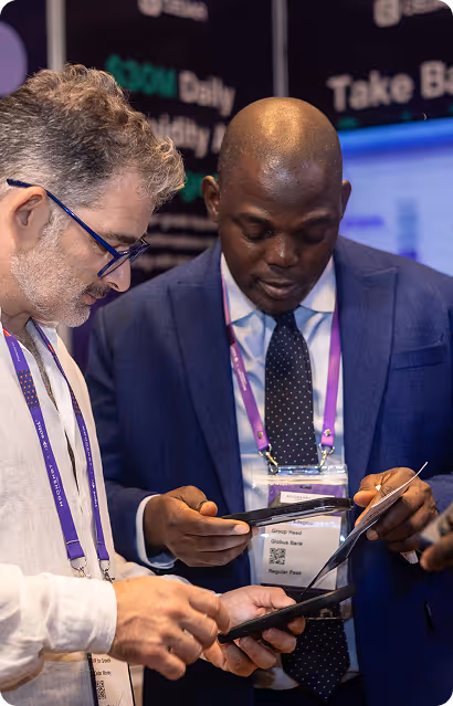 Two men wearing conference badges and lanyards looking at their smartphones, one in a blue suit and polka dot tie, the other in a white shirt and glasses.