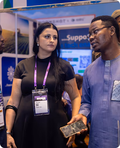 A woman with long dark hair wearing a black dress and conference badge listens to a man in glasses holding a phone at an event booth.