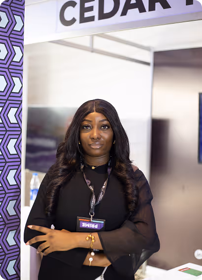 Woman with long curly hair wearing black attire and a conference badge standing with crossed arms in front of a display booth.