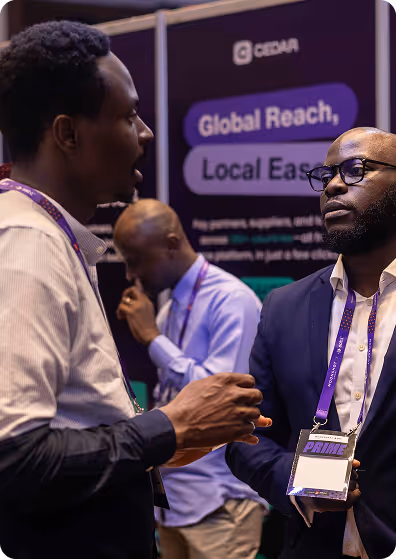 Two men engaged in conversation at a conference booth with a 'Global Reach, Local Ease' sign in the background, and a third man in the background looking at his phone.