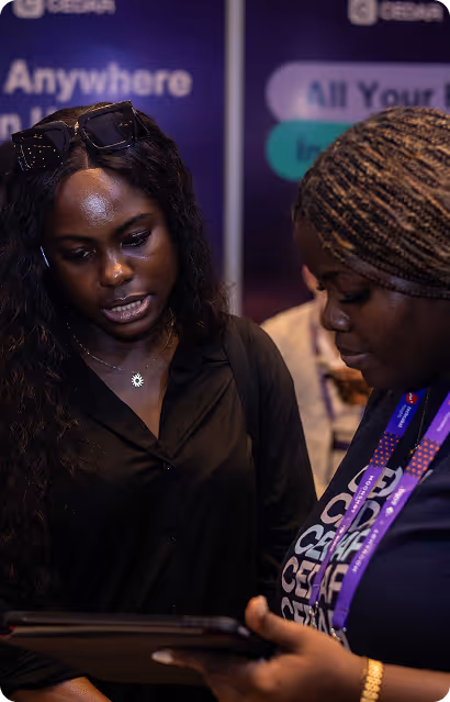Two women looking at a tablet, one wearing sunglasses on her head and the other with braided hair and a lanyard.