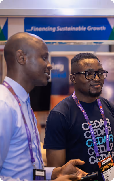 Two men wearing purple lanyards conversing at an event booth with a sign reading '...Financing Sustainable Growth' in the background.