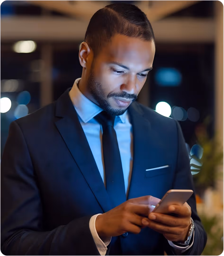 Man in a dark suit and tie looking at his smartphone in a dimly lit indoor setting at night.