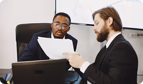 Two businessmen in suits reviewing documents together at a desk with a laptop.