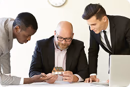 Three men in business attire examine a small house model at a table with a laptop.