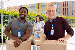 Two men smiling outdoors with ID badges, handling cardboard boxes, with a woman working behind them.