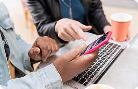 Two people using a smartphone together over a laptop keyboard, one pointing at the phone screen.