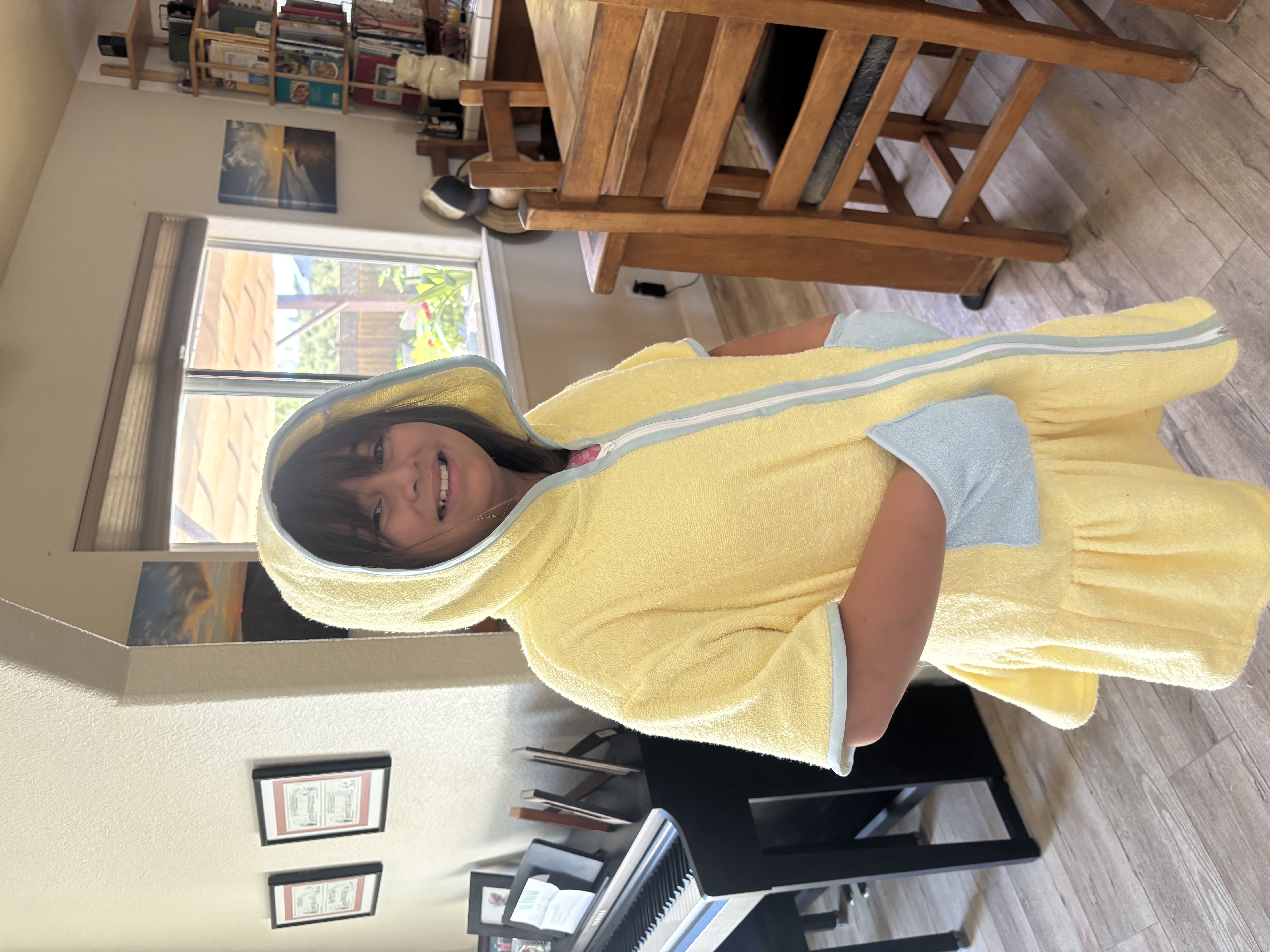 Smiling girl wearing a yellow hoodie towel with blue pockets standing indoors near a piano and wooden furniture.
